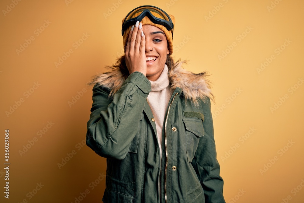 Young african american afro skier girl wearing snow sportswear and ski goggles covering one eye with hand, confident smile on face and surprise emotion.