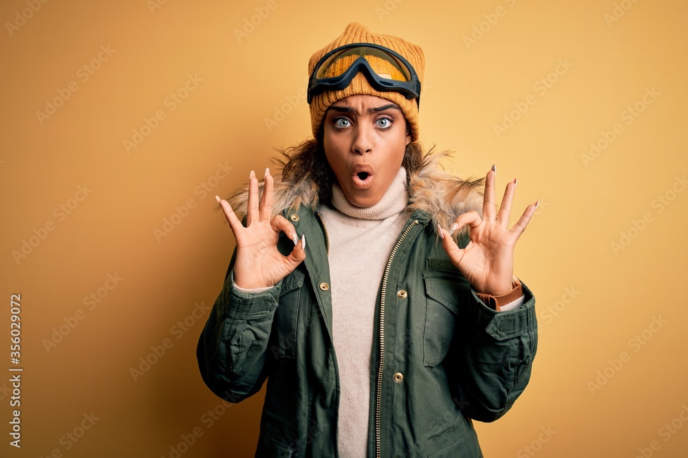 Young african american afro skier girl wearing snow sportswear and ski goggles looking surprised and shocked doing ok approval symbol with fingers. Crazy expression Young african american afro skier girl wearing snow sportswear and ski goggles looking surprised and shocked doing ok approval symbol with fingers. Crazy expression
