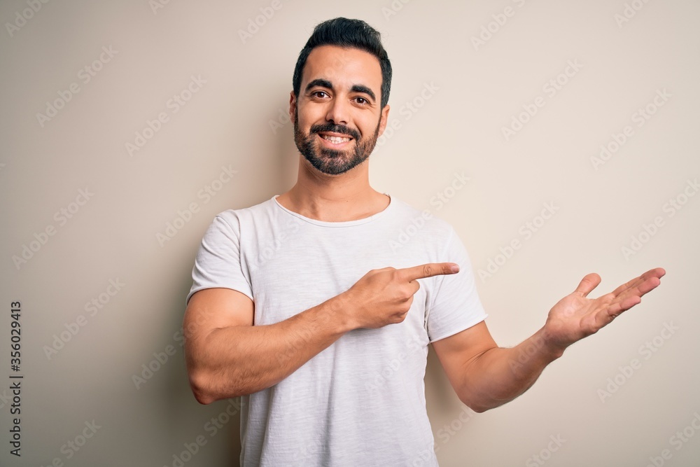 Young handsome man with beard wearing casual t-shirt standing over white background amazed and smiling to the camera while presenting with hand and pointing with finger. Young handsome man with beard wearing casual t-shirt standing over white background amazed and smiling to the camera while presenting with hand and pointing with finger.