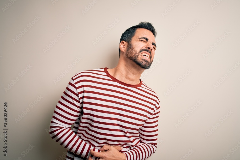 Young handsome man with beard wearing casual striped t-shirt standing over white background with hand on stomach because nausea, painful disease feeling unwell. Ache concept. Young handsome man with beard wearing casual striped t-shirt standing over white background with hand on stomach because nausea, painful disease feeling unwell. Ache concept.