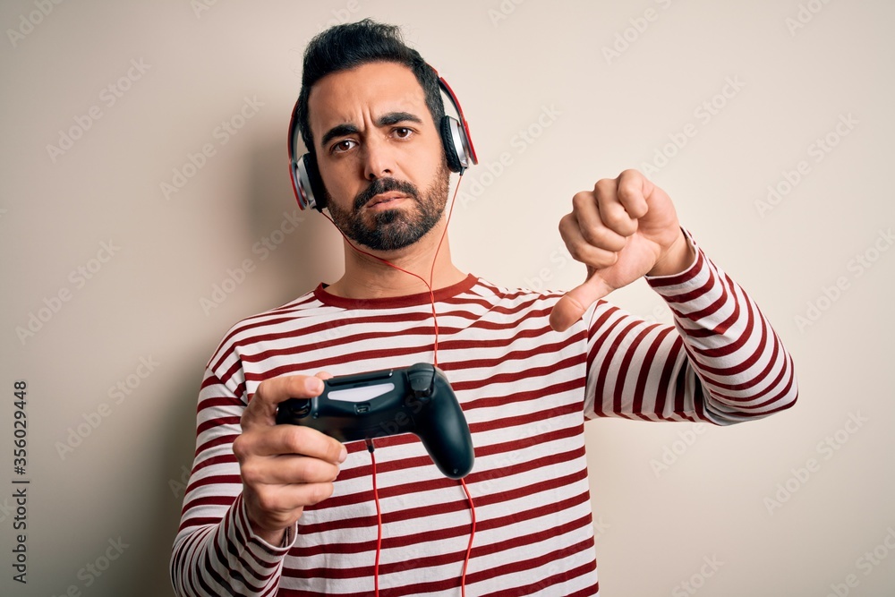 Young handsome gamer man with beard playing video game using joystick and headphones with angry face, negative sign showing dislike with thumbs down, rejection concept Young handsome gamer man with beard playing video game using joystick and headphones with angry face, negative sign showing dislike with thumbs down, rejection concept