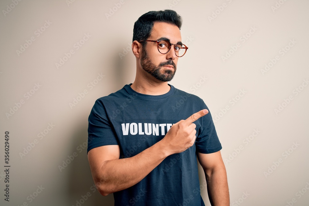 Handsome man with beard wearing t-shirt with volunteer message over white background Pointing with hand finger to the side showing advertisement, serious and calm face Handsome man with beard wearing t-shirt with volunteer message over white background Pointing with hand finger to the side showing advertisement, serious and calm face