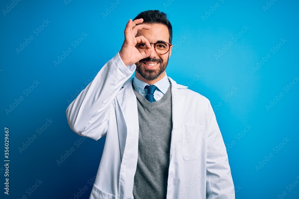 Young handsome doctor man with beard wearing coat and glasses over blue background doing ok gesture with hand smiling, eye looking through fingers with happy face. Young handsome doctor man with beard wearing coat and glasses over blue background doing ok gesture with hand smiling, eye looking through fingers with happy face.
