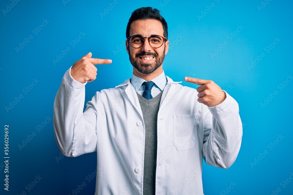 Young handsome doctor man with beard wearing coat and glasses over blue background smiling cheerful showing and pointing with fingers teeth and mouth. Dental health concept. Young handsome doctor man with beard wearing coat and glasses over blue background smiling cheerful showing and pointing with fingers teeth and mouth. Dental health concept.