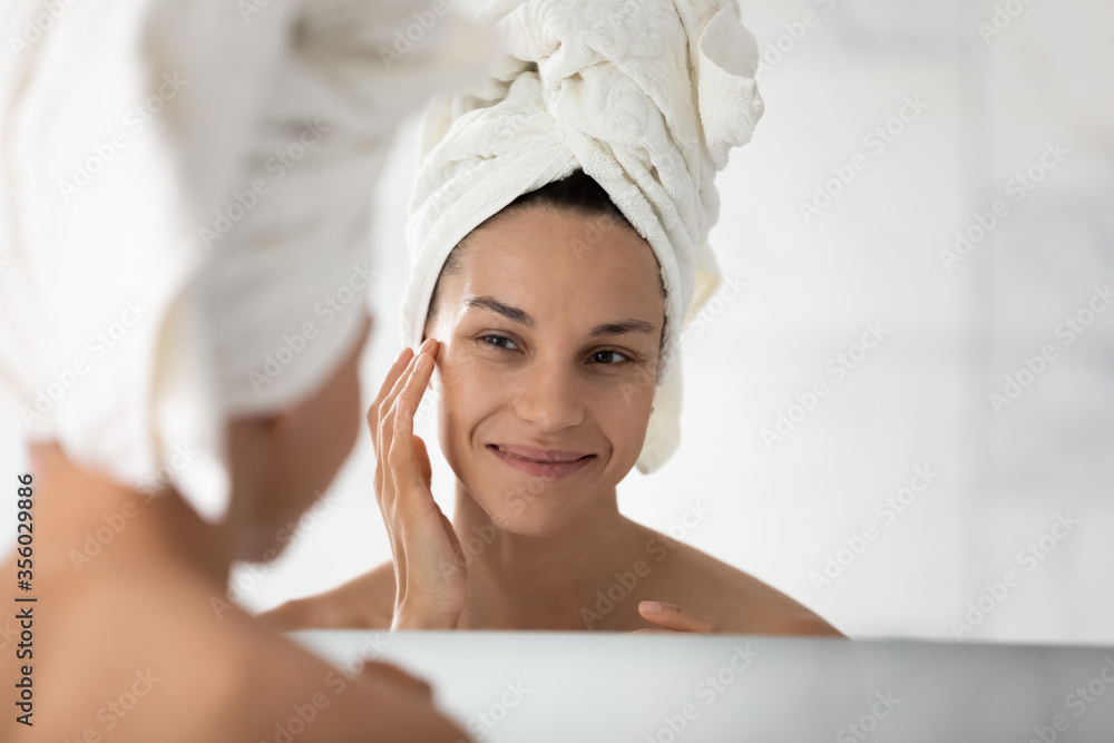 Head shot happy beautiful woman wearing white bath towel on head touching perfect healthy skin, standing in bathroom, looking in mirror, doing face massage or applying moisturizing face cream Head shot happy beautiful woman wearing white bath towel on head touching perfect healthy skin, standing in bathroom, looking in mirror, doing face massage or applying moisturizing face cream