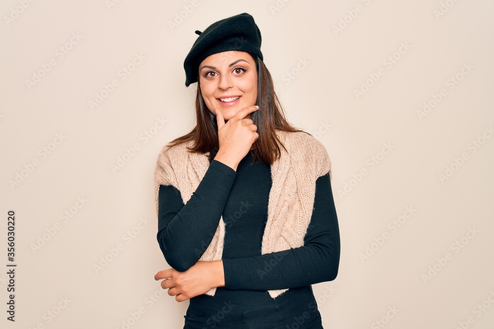 Young beautiful brunette woman wearing casual french beret over white background looking confident at the camera smiling with crossed arms and hand raised on chin. Thinking positive. Young beautiful brunette woman wearing casual french beret over white background looking confident at the camera smiling with crossed arms and hand raised on chin. Thinking positive.