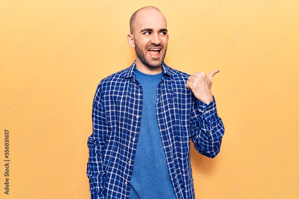 Young handsome bald man wearing casual shirt pointing thumb up to the side smiling happy with open mouth Young handsome bald man wearing casual shirt pointing thumb up to the side smiling happy with open mouth
