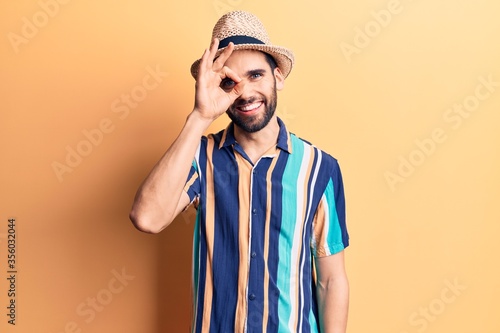 Young handsome man with beard wearing summer hat and shirt doing ok gesture with hand smiling, eye looking through fingers with happy face.
