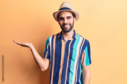 Young handsome man with beard wearing summer hat and shirt smiling cheerful presenting and pointing with palm of hand looking at the camera.