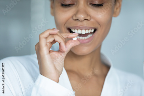 Close up smiling African American young woman holding white round pill, beautiful girl wearing white bathrobe taking supplements or vitamins in morning, healthcare and treatment concept