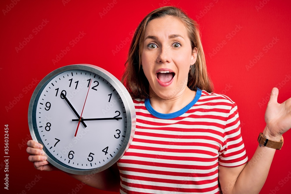 Young beautiful blonde woman holding big clock standing over isolated red background very happy and excited, winner expression celebrating victory screaming with big smile and raised hands Young beautiful blonde woman holding big clock standing over isolated red background very happy and excited, winner expression celebrating victory screaming with big smile and raised hands