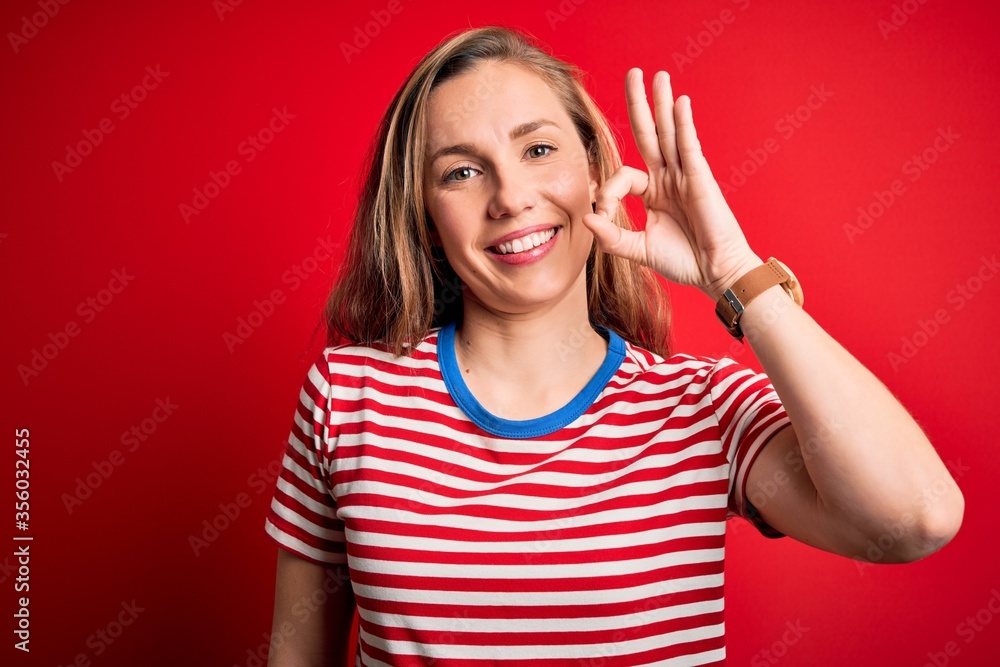 Fototapeta premium Young beautiful blonde woman wearing casual striped t-shirt over isolated red background smiling positive doing ok sign with hand and fingers. Successful expression.