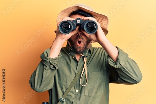 Young hispanic man wearing explorer hat holding binoculars scared and amazed with open mouth for surprise, disbelief face photo