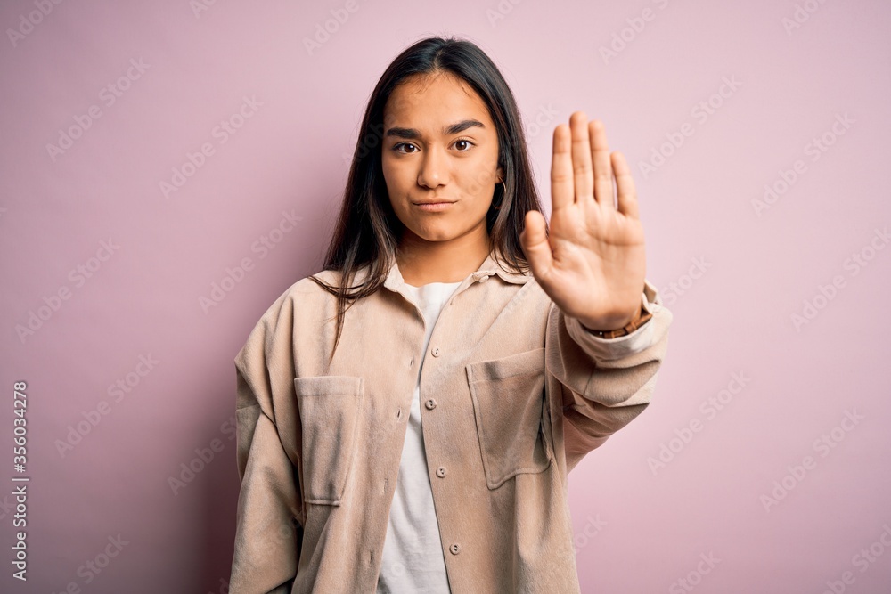 Young beautiful asian woman wearing casual shirt standing over pink background doing stop sing with palm of the hand. Warning expression with negative and serious gesture on the face.