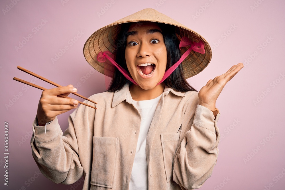 Young thai woman wearing traditional conical asian hat eating food using chopsticks very happy and excited, winner expression celebrating victory screaming with big smile and raised hands Young thai woman wearing traditional conical asian hat eating food using chopsticks very happy and excited, winner expression celebrating victory screaming with big smile and raised hands