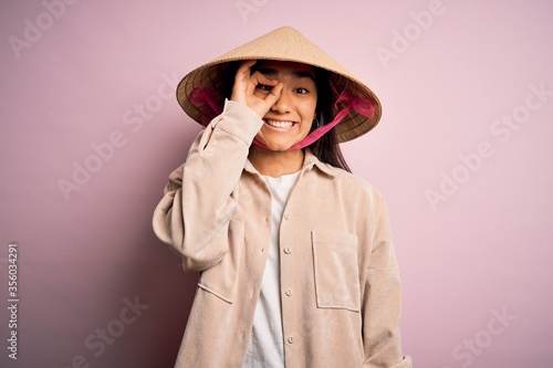 Young beautiful woman wearing traditional conical asian hat over isolated pink background doing ok gesture with hand smiling, eye looking through fingers with happy face.