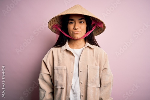Young beautiful woman wearing traditional conical asian hat over isolated pink background with serious expression on face. Simple and natural looking at the camera.