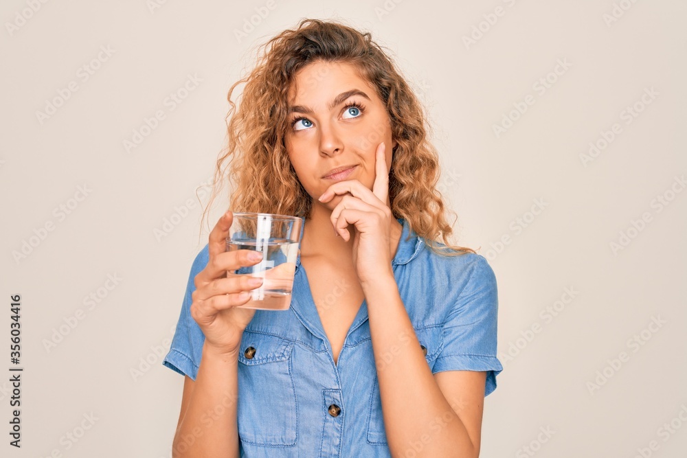 Young beautiful blonde woman with blue eyes drinking glass of water over white background serious face thinking about question, very confused idea Young beautiful blonde woman with blue eyes drinking glass of water over white background serious face thinking about question, very confused idea