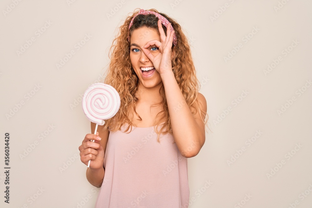 Young beautiful blonde woman with blue eyes eating sweet candy lollipop over pink background with happy face smiling doing ok sign with hand on eye looking through fingers Young beautiful blonde woman with blue eyes eating sweet candy lollipop over pink background with happy face smiling doing ok sign with hand on eye looking through fingers