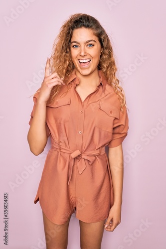 Young beautiful woman with blue eyes wearing casual summer dress over pink background pointing finger up with successful idea. Exited and happy. Number one.