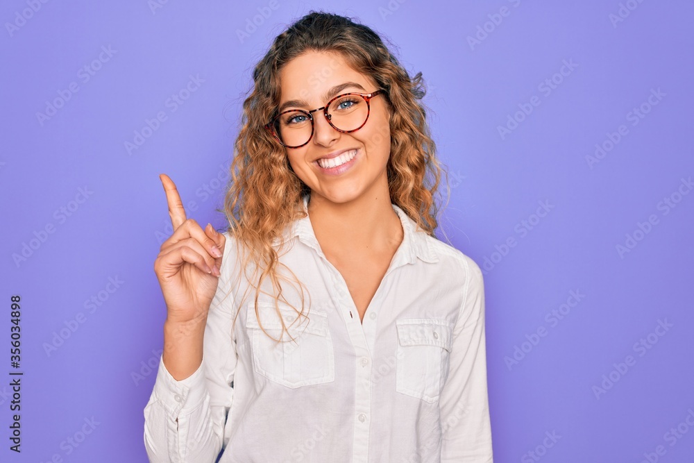 Young beautiful woman with blue eyes wearing casual shirt and glasses over purple background showing and pointing up with finger number one while smiling confident and happy. Young beautiful woman with blue eyes wearing casual shirt and glasses over purple background showing and pointing up with finger number one while smiling confident and happy.