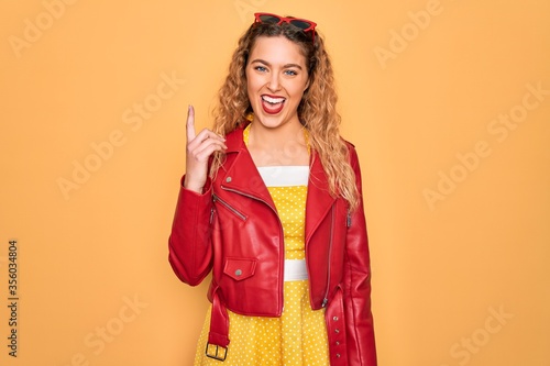 Young beautiful blonde woman pin-up with blue eyes wearing red sunglasses and jacket pointing finger up with successful idea. Exited and happy. Number one.