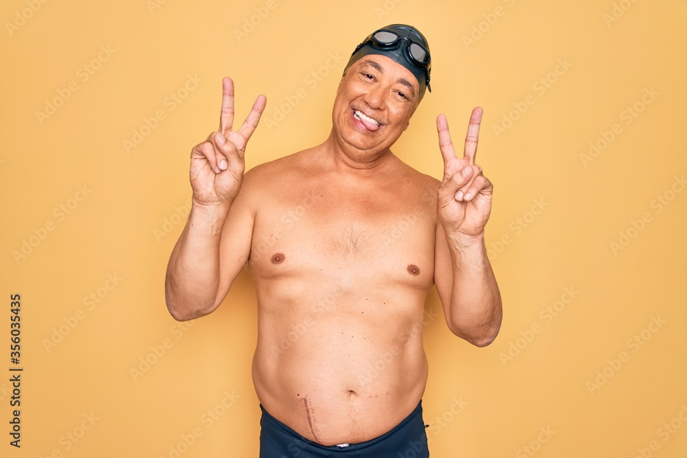Middle age senior grey-haired swimmer man wearing swimsuit, cap and goggles smiling with tongue out showing fingers of both hands doing victory sign. Number two.