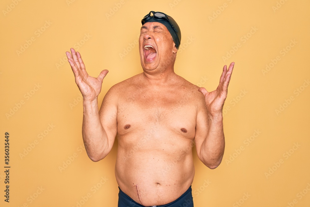 Middle age senior grey-haired swimmer man wearing swimsuit, cap and goggles celebrating mad and crazy for success with arms raised and closed eyes screaming excited. Winner concept
