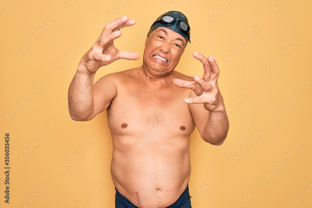 Middle age senior grey-haired swimmer man wearing swimsuit, cap and goggles Shouting frustrated with rage, hands trying to strangle, yelling mad