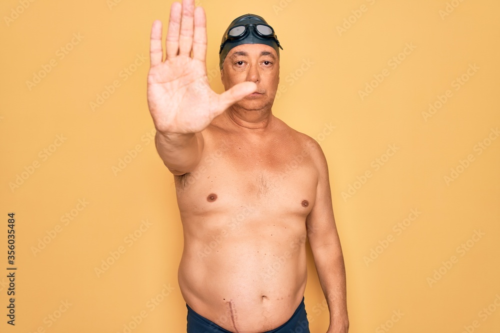 Middle age senior grey-haired swimmer man wearing swimsuit, cap and goggles doing stop sing with palm of the hand. Warning expression with negative and serious gesture on the face.