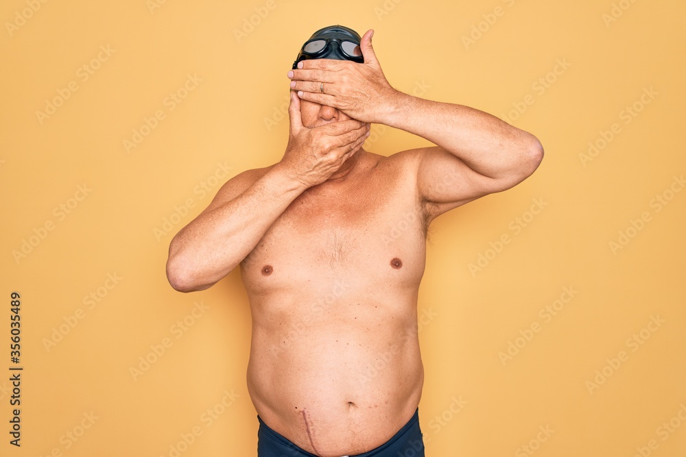 Middle age senior grey-haired swimmer man wearing swimsuit, cap and goggles Covering eyes and mouth with hands, surprised and shocked. Hiding emotion