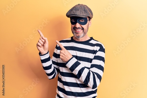 Middle age handsome burglar man wearing cap and mask over isolated yellow background smiling and looking at the camera pointing with two hands and fingers to the side. © Krakenimages.com