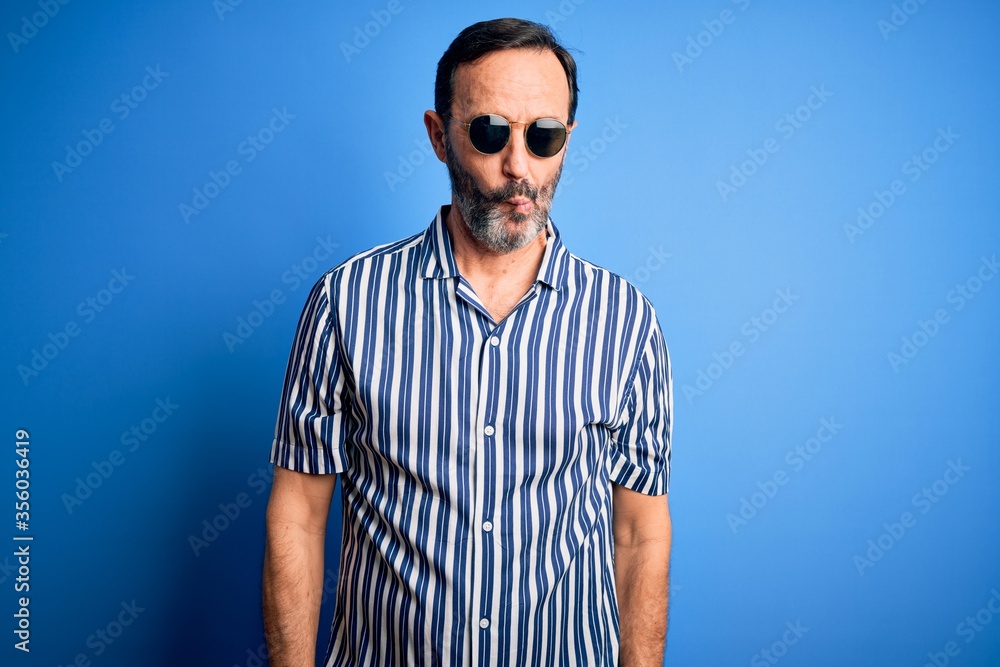 Middle age hoary man wearing striped shirt and sunglasses over isolated blue background making fish face with lips, crazy and comical gesture. Funny expression. Middle age hoary man wearing striped shirt and sunglasses over isolated blue background making fish face with lips, crazy and comical gesture. Funny expression.