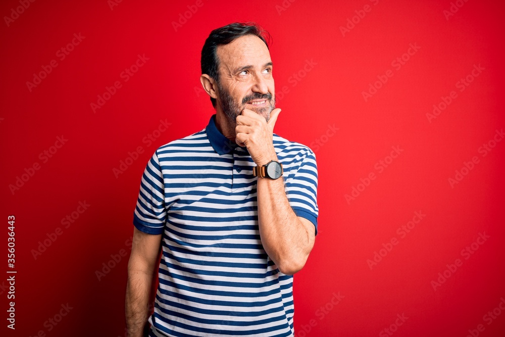 Middle age hoary man wearing casual striped polo standing over isolated red background with hand on chin thinking about question, pensive expression. Smiling with thoughtful face. Doubt concept.