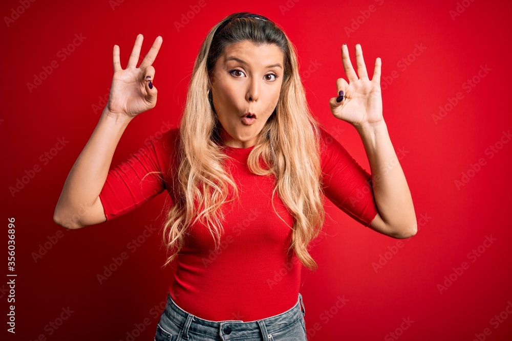 Young beautiful blonde woman wearing casual t-shirt standing over isolated red background looking surprised and shocked doing ok approval symbol with fingers. Crazy expression Young beautiful blonde woman wearing casual t-shirt standing over isolated red background looking surprised and shocked doing ok approval symbol with fingers. Crazy expression