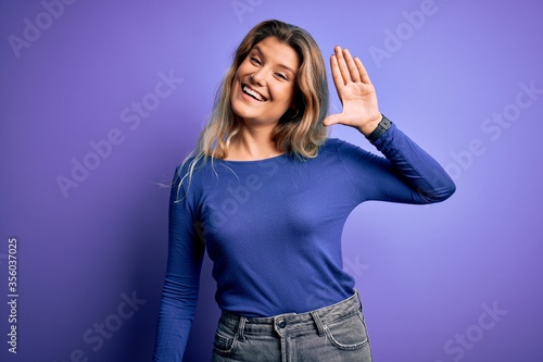 Young beautiful blonde woman wearing casual t-shirt over isolated purple background Waiving saying hello happy and smiling, friendly welcome gesture