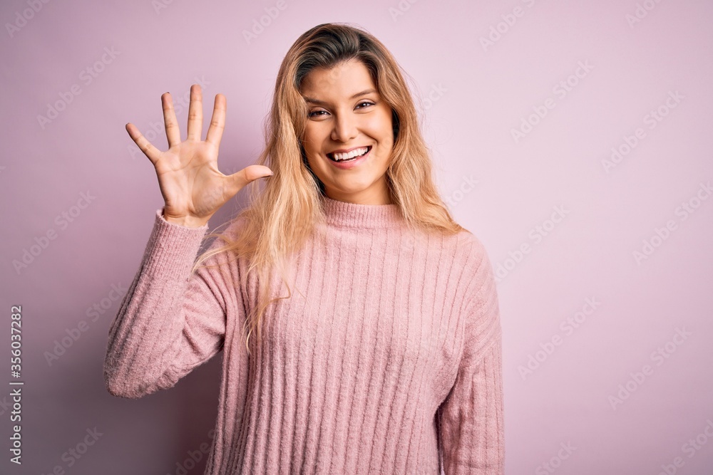 Young beautiful blonde woman wearing casual pink sweater over isolated background showing and pointing up with fingers number five while smiling confident and happy. Young beautiful blonde woman wearing casual pink sweater over isolated background showing and pointing up with fingers number five while smiling confident and happy.