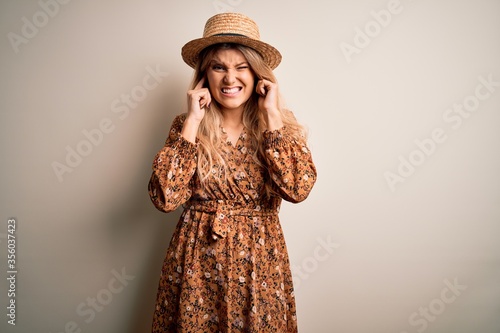 Young beautiful blonde woman wearing summer dress and hat over isolated white background covering ears with fingers with annoyed expression for the noise of loud music. Deaf concept.