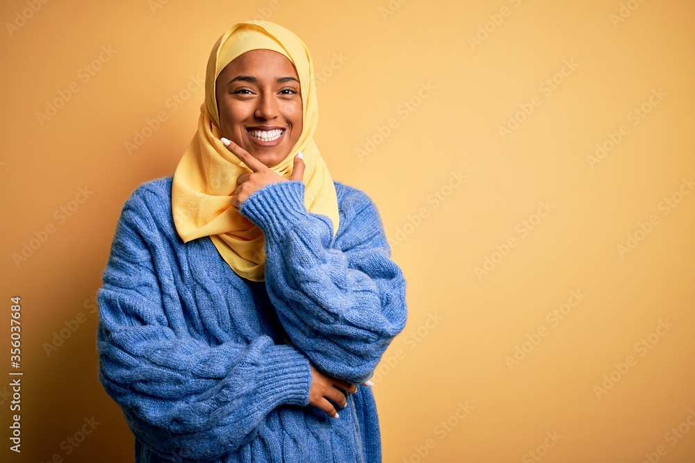Young African American afro woman wearing muslim hijab over isolated yellow background looking confident at the camera smiling with crossed arms and hand raised on chin. Thinking positive. Young African American afro woman wearing muslim hijab over isolated yellow background looking confident at the camera smiling with crossed arms and hand raised on chin. Thinking positive.