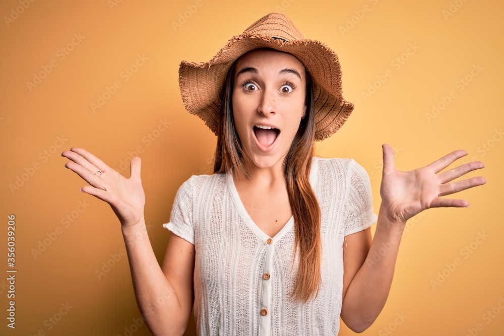Young beautiful woman wearing casual t-shirt and summer hat over isolated yellow background celebrating crazy and amazed for success with arms raised and open eyes screaming excited. Winner concept Young beautiful woman wearing casual t-shirt and summer hat over isolated yellow background celebrating crazy and amazed for success with arms raised and open eyes screaming excited. Winner concept