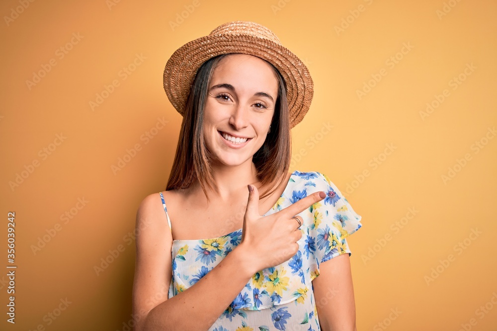 Young beautiful woman wearing casual t-shirt and summer hat over isolated yellow background cheerful with a smile of face pointing with hand and finger up to the side with happy and natural expression