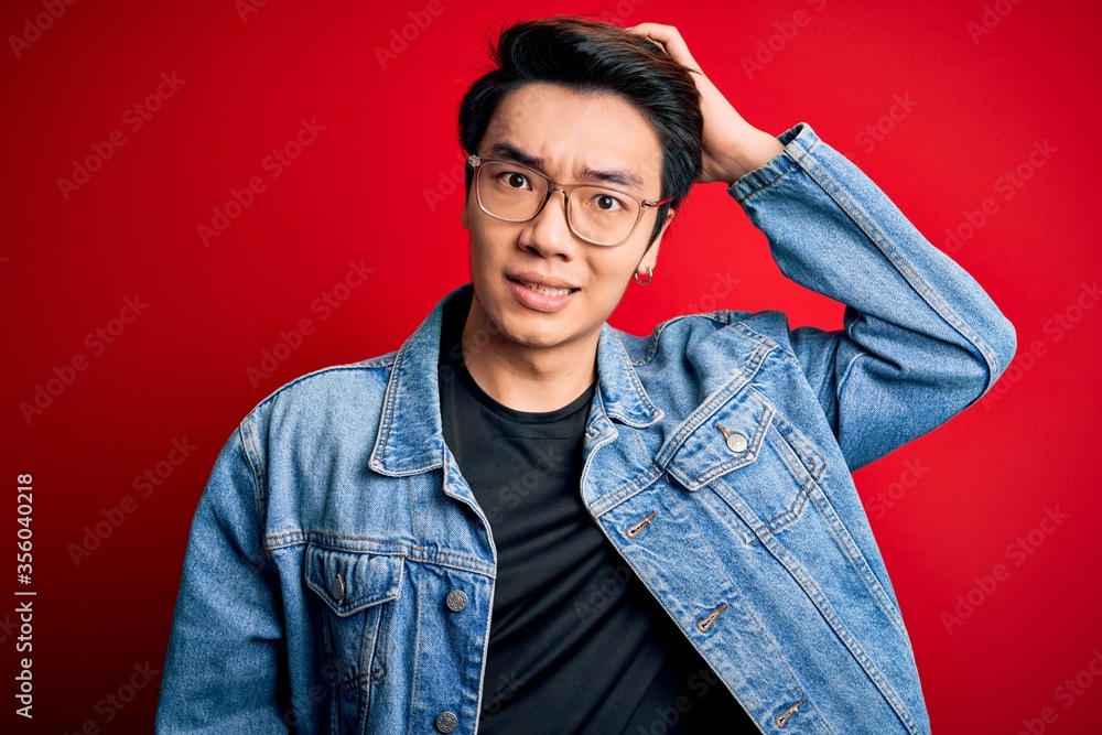 Young handsome chinese man wearing denim jacket and glasses over red background confuse and wonder about question. Uncertain with doubt, thinking with hand on head. Pensive concept. Young handsome chinese man wearing denim jacket and glasses over red background confuse and wonder about question. Uncertain with doubt, thinking with hand on head. Pensive concept.