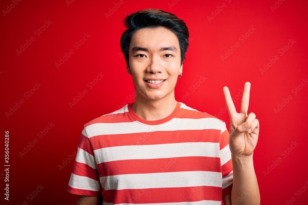 Young handsome chinese man wearing casual striped t-shirt standing over red background showing and pointing up with fingers number two while smiling confident and happy. Young handsome chinese man wearing casual striped t-shirt standing over red background showing and pointing up with fingers number two while smiling confident and happy.