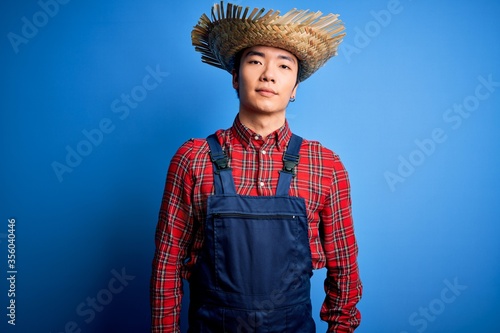 Young handsome chinese farmer man wearing apron and straw hat over blue background Relaxed with serious expression on face. Simple and natural looking at the camera.