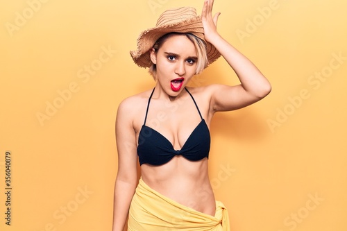 Young beautiful blonde woman wearing bikini and summer hat surprised with hand on head for mistake  remember error. forgot  bad memory concept.