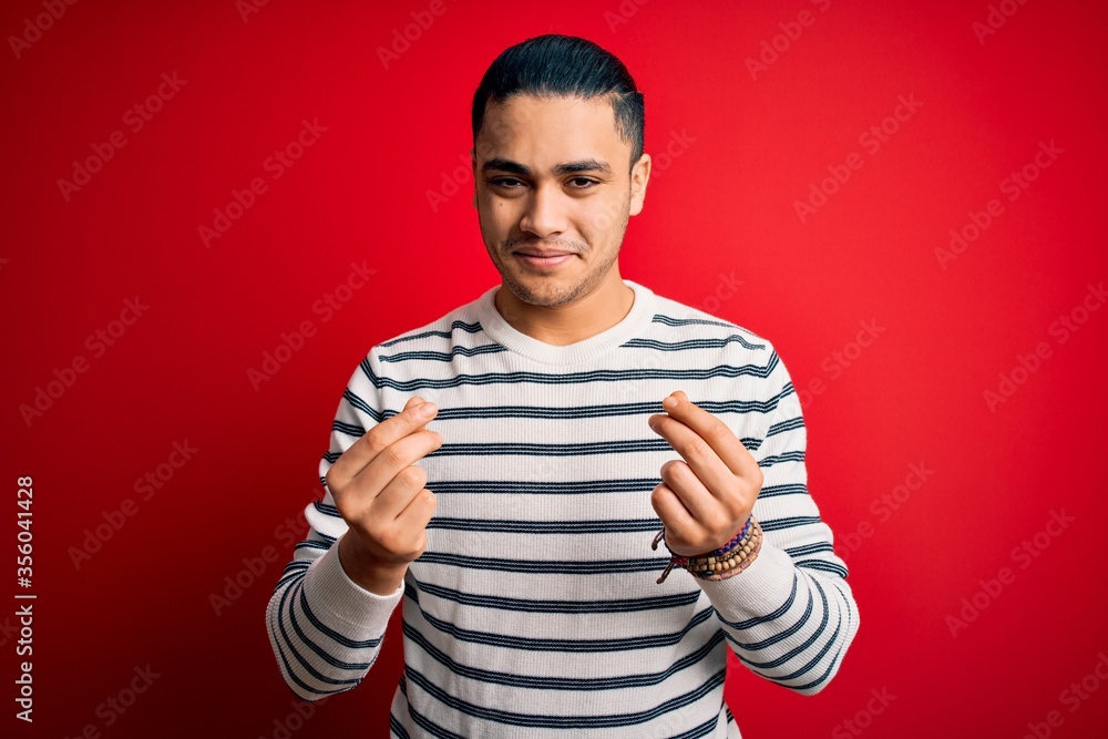 Young brazilian man wearing casual striped t-shirt standing over isolated red background doing money gesture with hands, asking for salary payment, millionaire business Young brazilian man wearing casual striped t-shirt standing over isolated red background doing money gesture with hands, asking for salary payment, millionaire business
