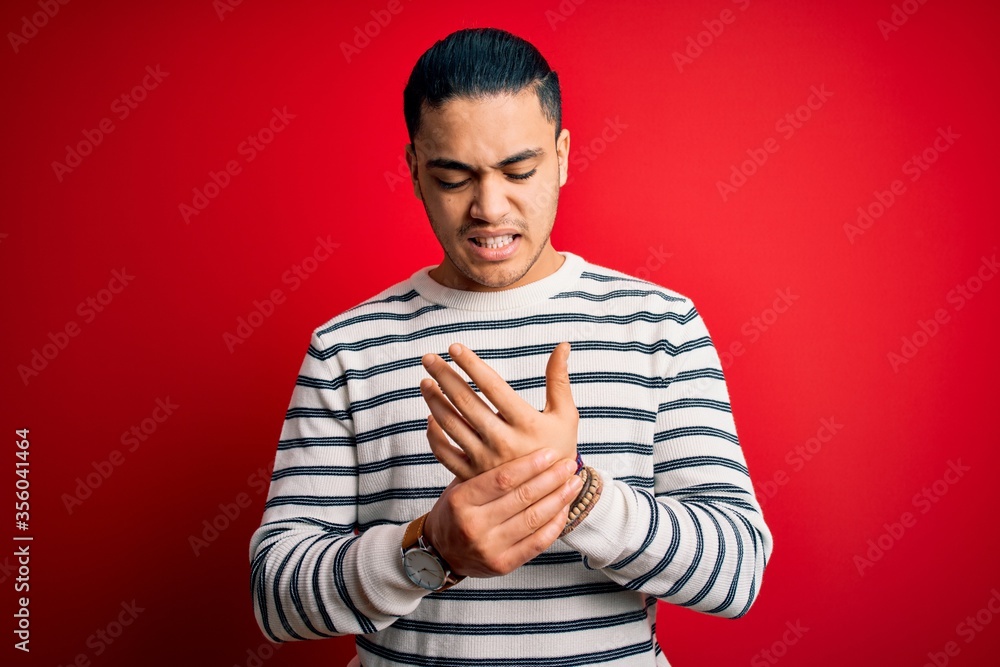 Young brazilian man wearing casual striped t-shirt standing over isolated red background Suffering pain on hands and fingers, arthritis inflammation Young brazilian man wearing casual striped t-shirt standing over isolated red background Suffering pain on hands and fingers, arthritis inflammation
