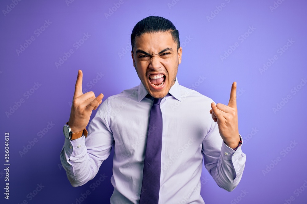 Young brazilian businessman wearing elegant tie standing over isolated purple background shouting with crazy expression doing rock symbol with hands up. Music star. Heavy music concept. Young brazilian businessman wearing elegant tie standing over isolated purple background shouting with crazy expression doing rock symbol with hands up. Music star. Heavy music concept.