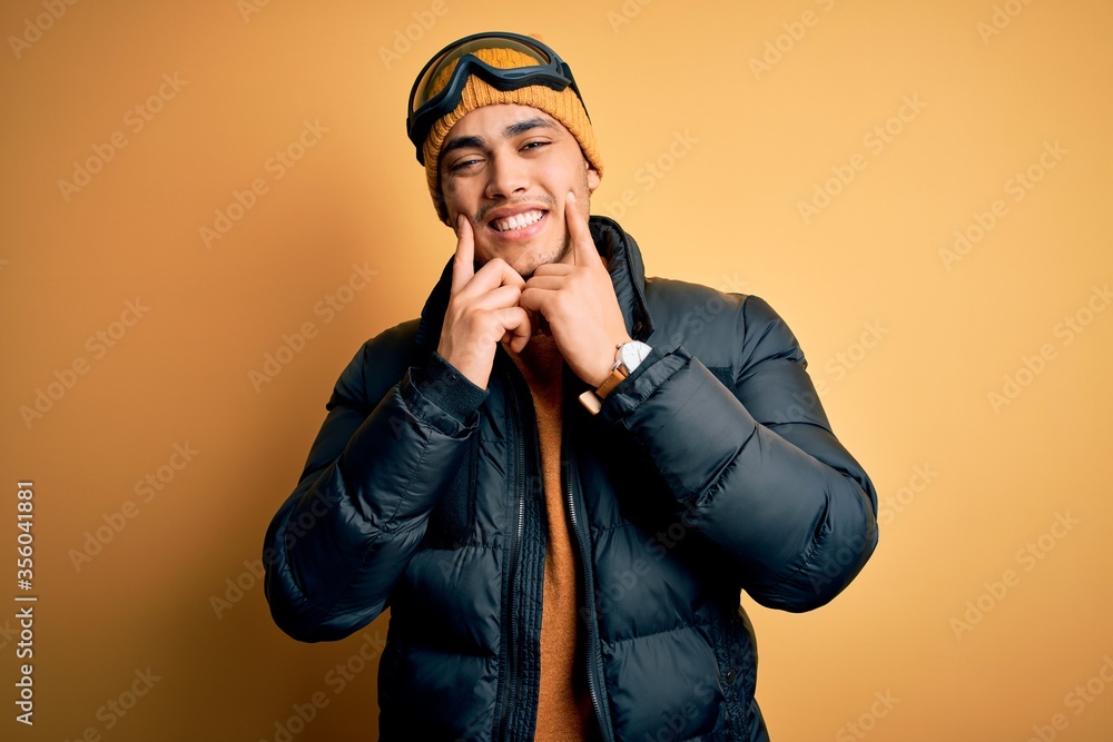 Young brazilian skier man wearing snow sportswear and ski goggles over yellow background Smiling with open mouth, fingers pointing and forcing cheerful smile Young brazilian skier man wearing snow sportswear and ski goggles over yellow background Smiling with open mouth, fingers pointing and forcing cheerful smile