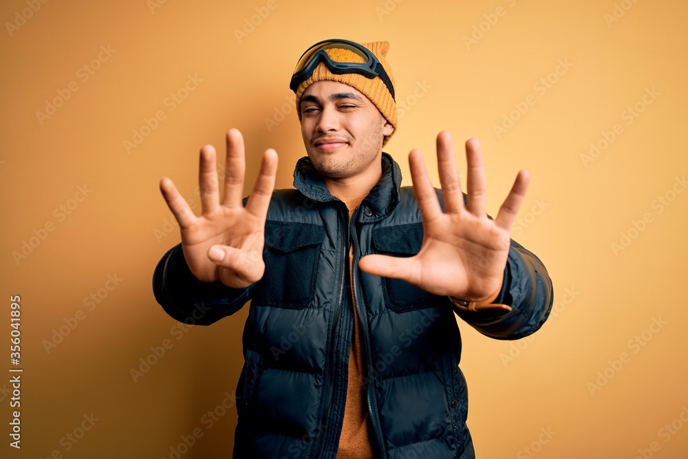 Young brazilian skier man wearing snow sportswear and ski goggles over yellow background showing and pointing up with fingers number nine while smiling confident and happy. Young brazilian skier man wearing snow sportswear and ski goggles over yellow background showing and pointing up with fingers number nine while smiling confident and happy.